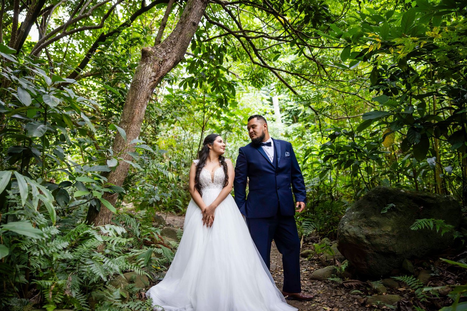 Bride in a white gown and groom in a navy suit stand together in a lush green tropical garden for a wedding portrait. Iao Valley