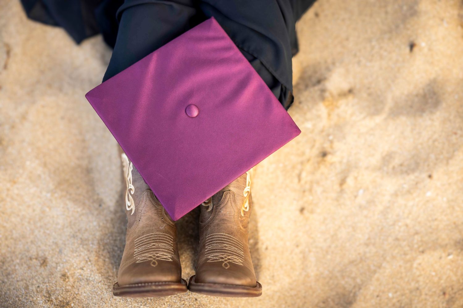 Purple graduation cap resting on the lap of a graduate in brown cowboy boots sitting on sandy ground. Paia
