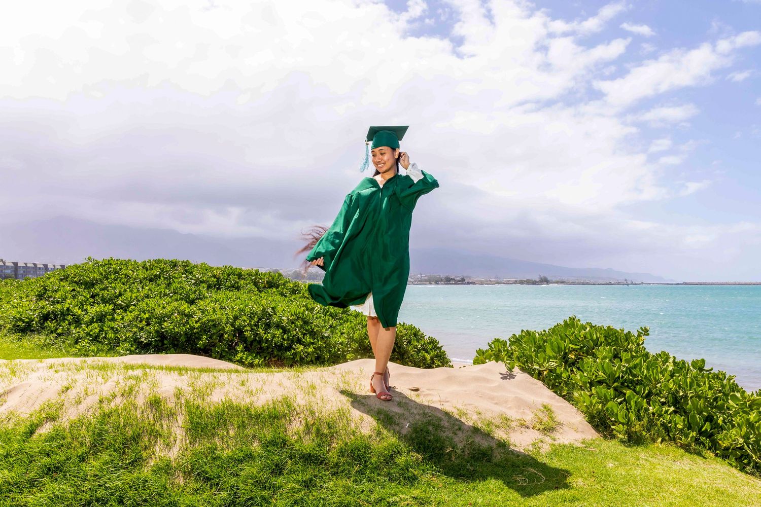 Graduate in a green cap and gown poses on a grassy coastal hill with turquoise ocean water and a cloudy blue sky behind. Kahului