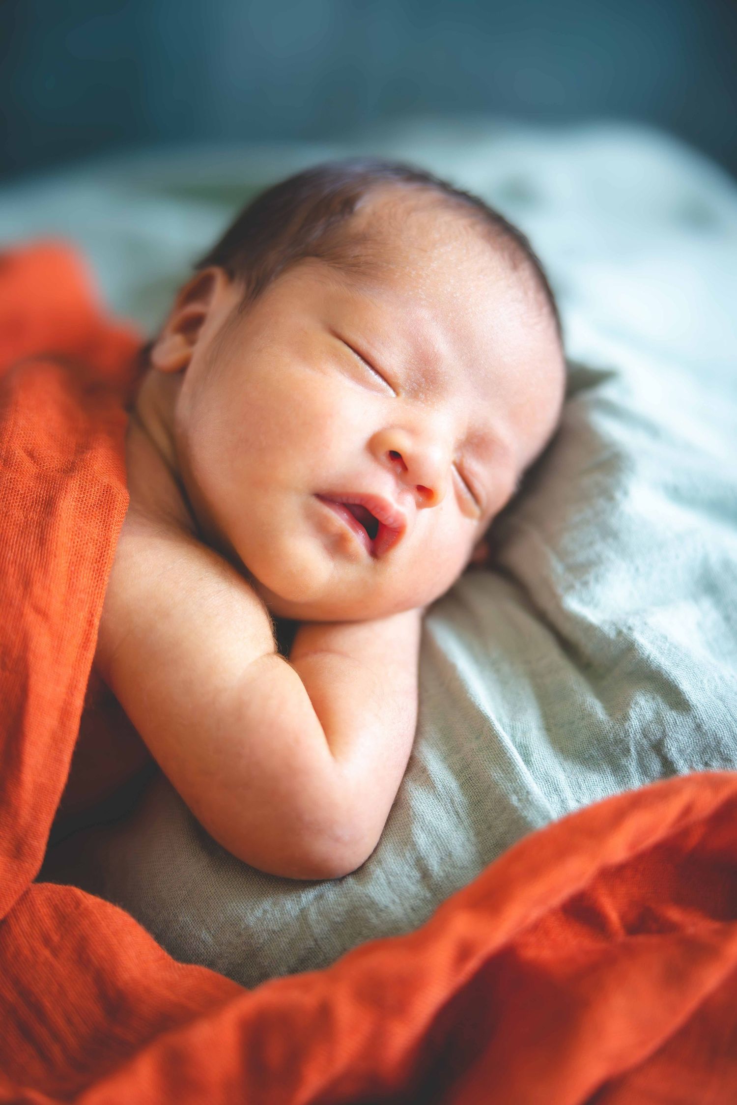 Peaceful newborn sleeps with hands tucked under its cheek, resting on a gray blanket beside a bright orange cloth.