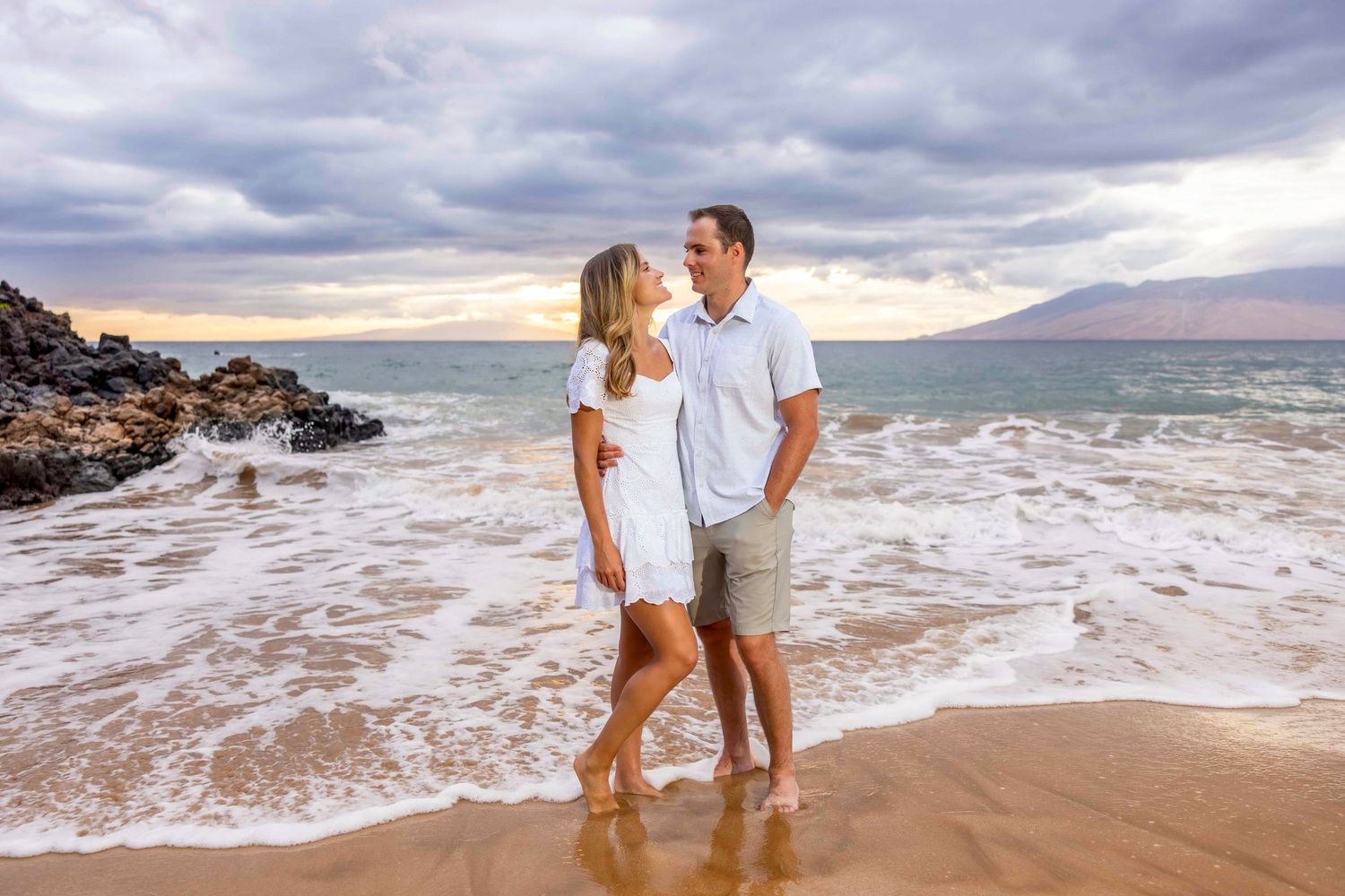 Smiling couple in white outfits stand barefoot on a sandy beach as ocean waves roll in beneath a dramatic cloudy sunset sky. Wailea