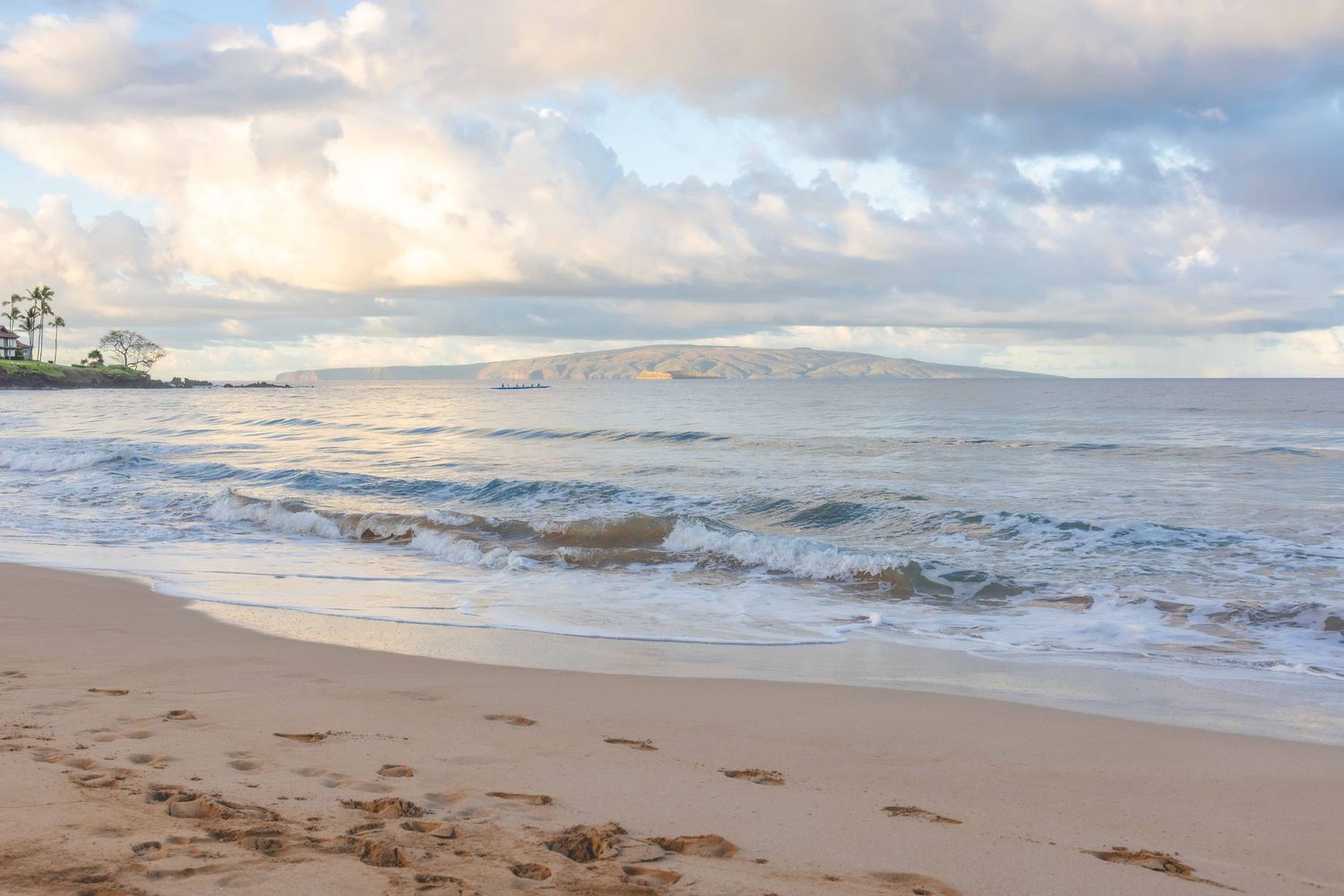 Gentle ocean waves roll onto a sandy tropical beach with footprints, distant island hills, and soft clouds across a pastel sky at sunset.
