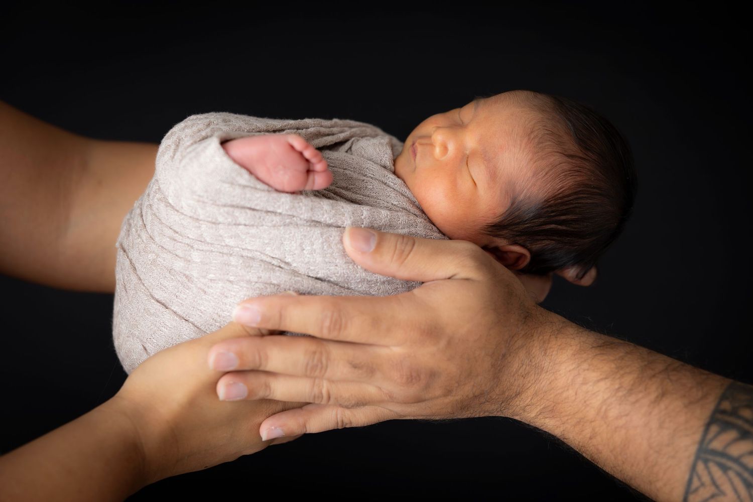 Sleeping newborn in a gray knit wrap cradled in a parent's hands against a black background in a studio portrait.