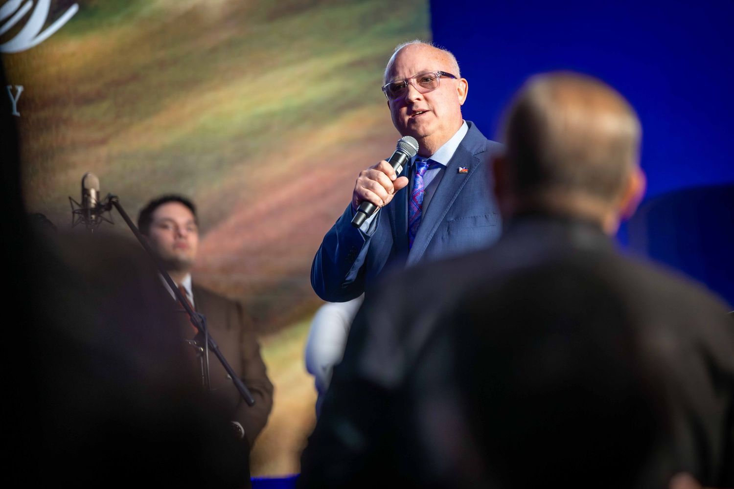 Gray-haired man in glasses and a blue suit speaks into a microphone on stage while audience members watch in the foreground.
