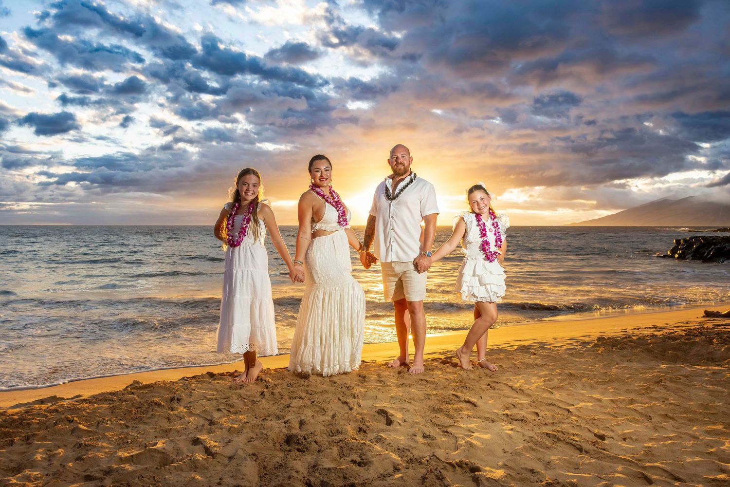 Family of four in white outfits and flower leis holding hands on a sandy beach at sunset with ocean waves and a colorful cloudy sky.