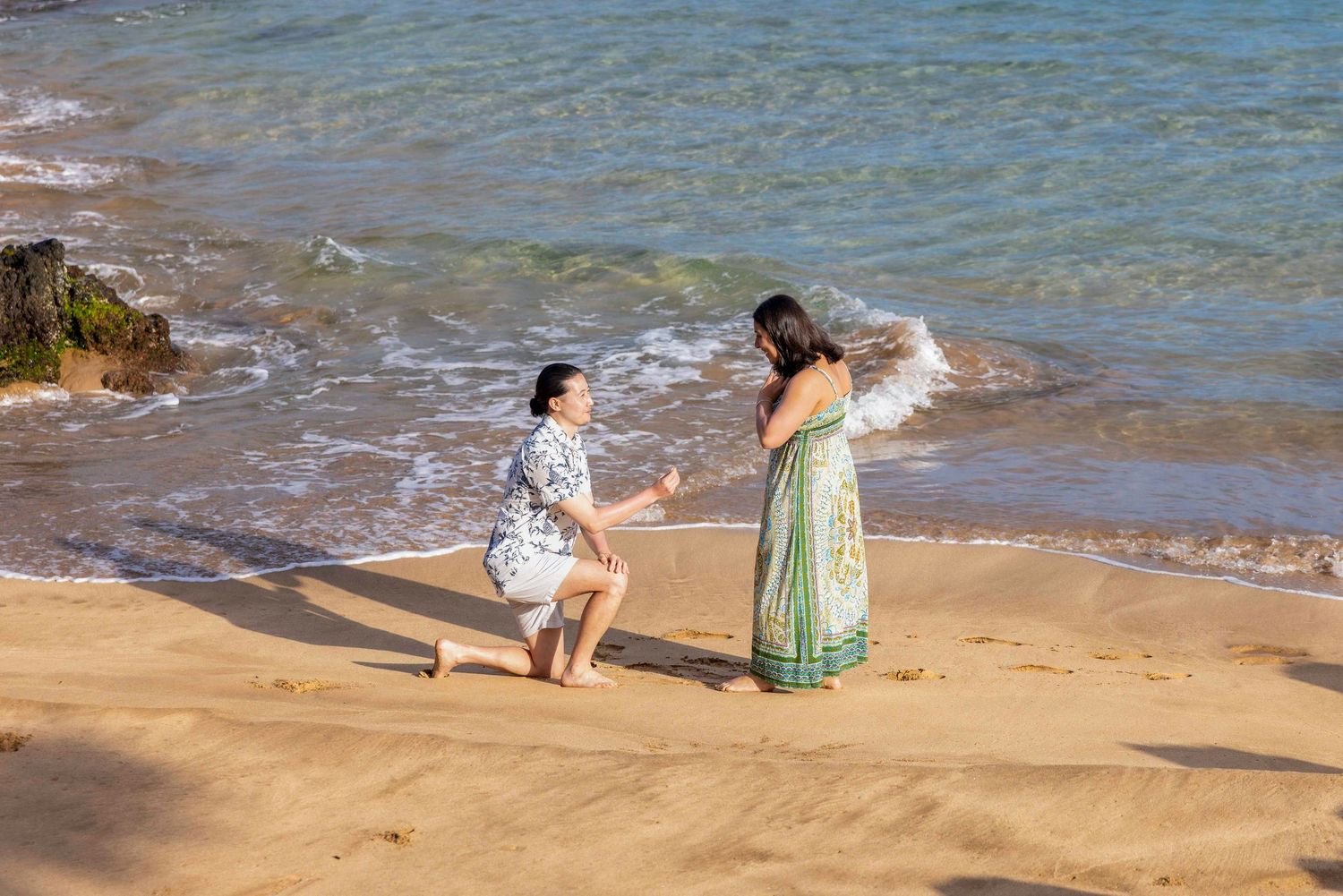 Man kneeling on one knee proposes to a woman in a green patterned dress on a sandy beach with gentle ocean waves in the background. Makena