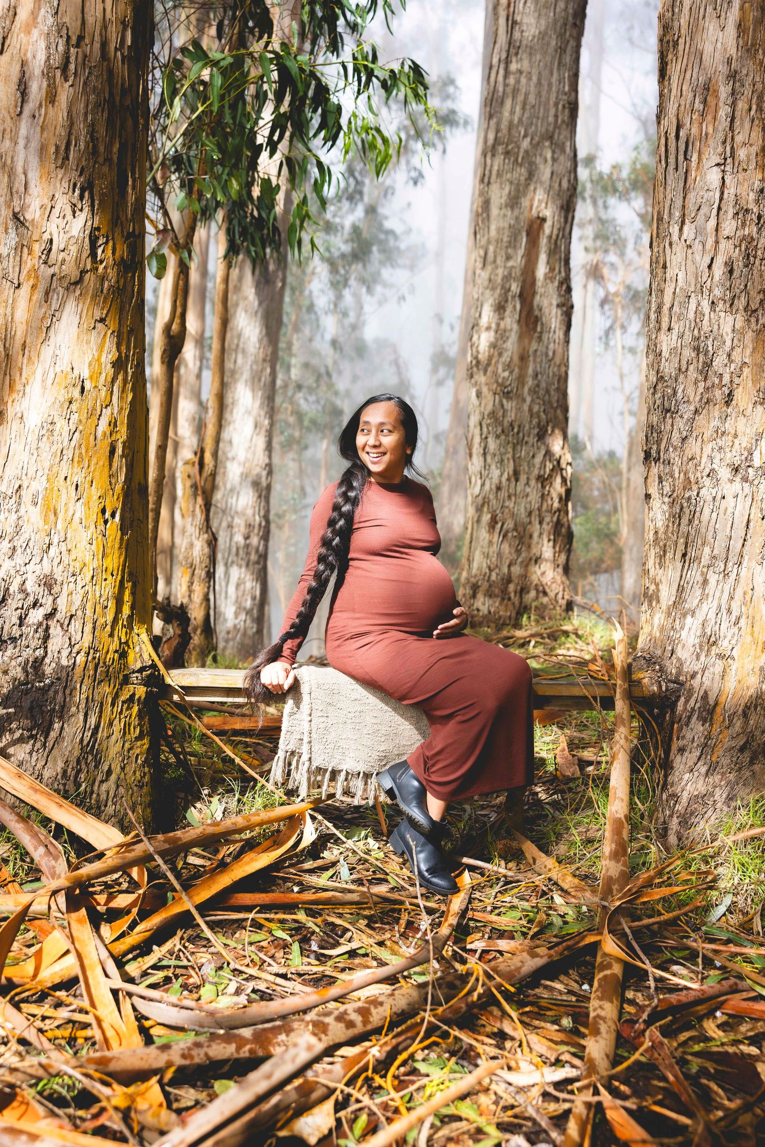 Pregnant woman in a fitted rust-colored dress sits on a wooden bench smiling among tall eucalyptus trees in a misty forest. Poli Poli