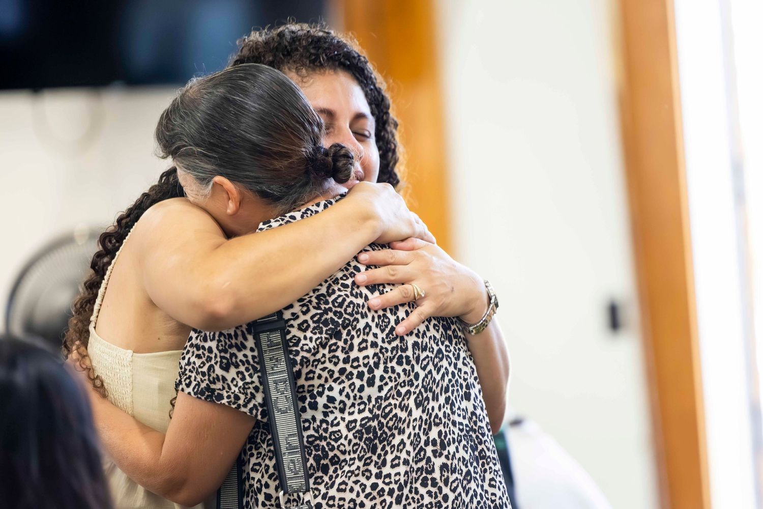 Two women share a warm, emotional hug in a bright room, one wearing a leopard print top with her dark hair pulled back.