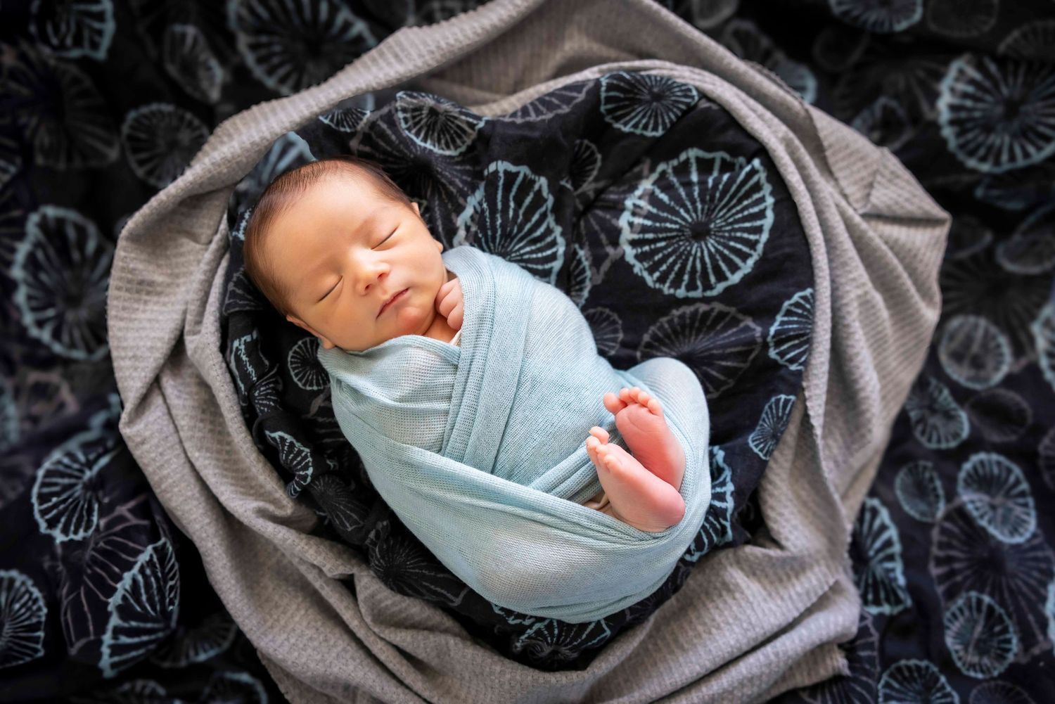 Newborn baby sleeping peacefully wrapped in a light blue swaddle on a gray blanket with a dark floral backdrop, bare feet peeking out.