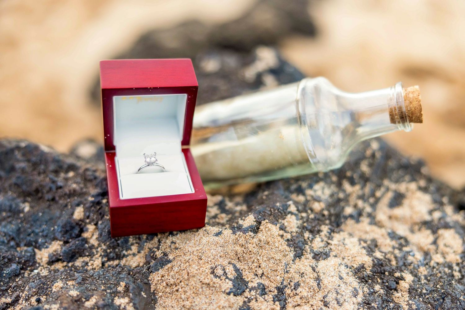 Red box holding a diamond engagement ring beside a corked glass message bottle on dark volcanic rock and sand at the beach.