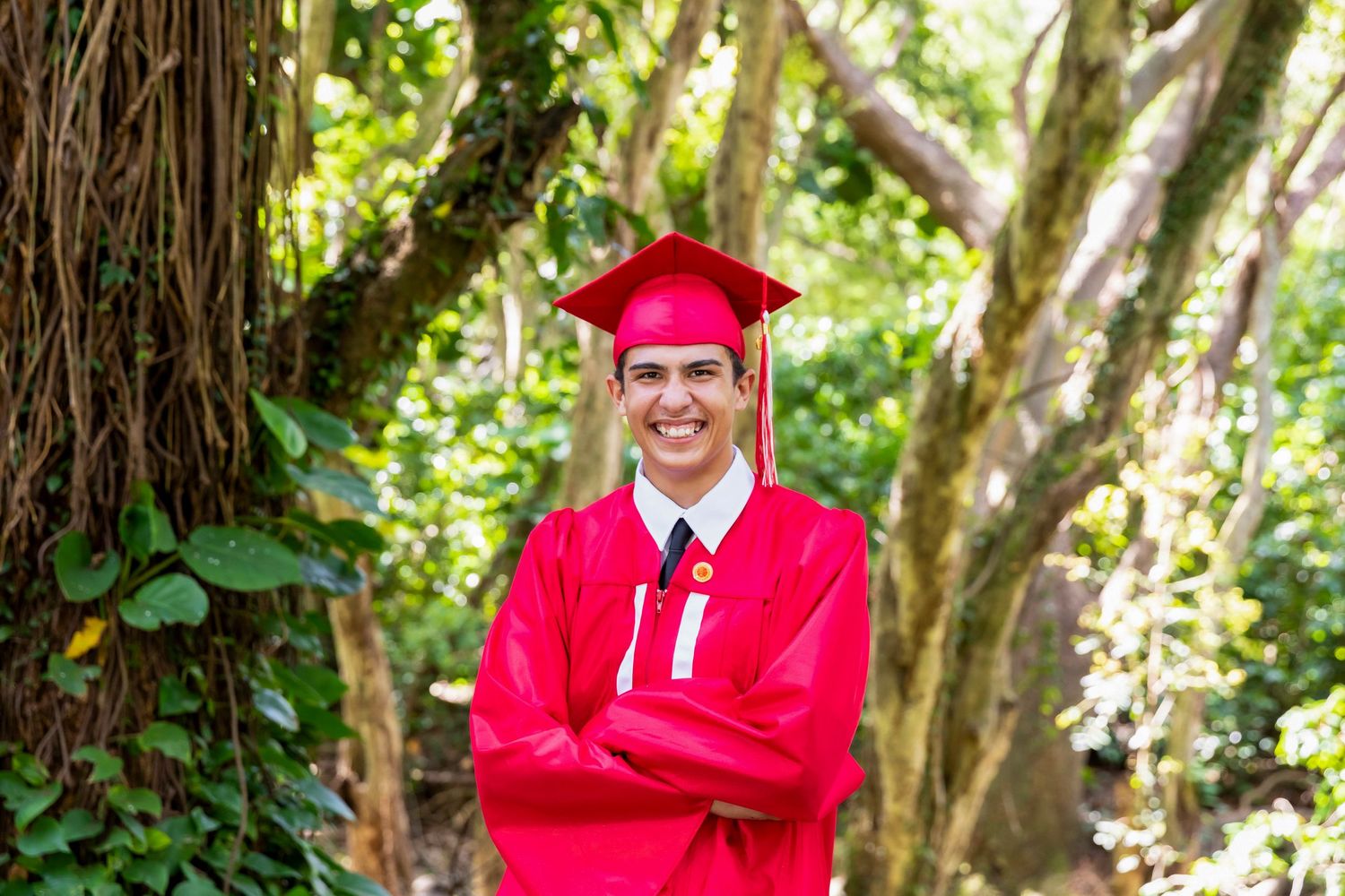 Smiling graduate in a red cap and gown stands with arms crossed among lush green trees and tropical foliage in an outdoor portrait. Honolua