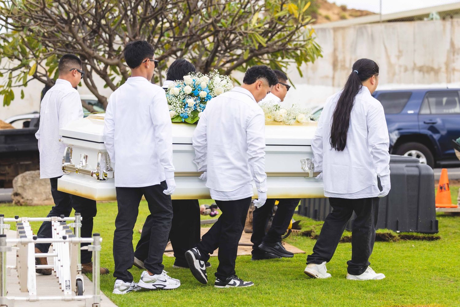 Pallbearers in white shirts and dark pants carry a white casket topped with flowers across a grassy cemetery lawn during a funeral service.