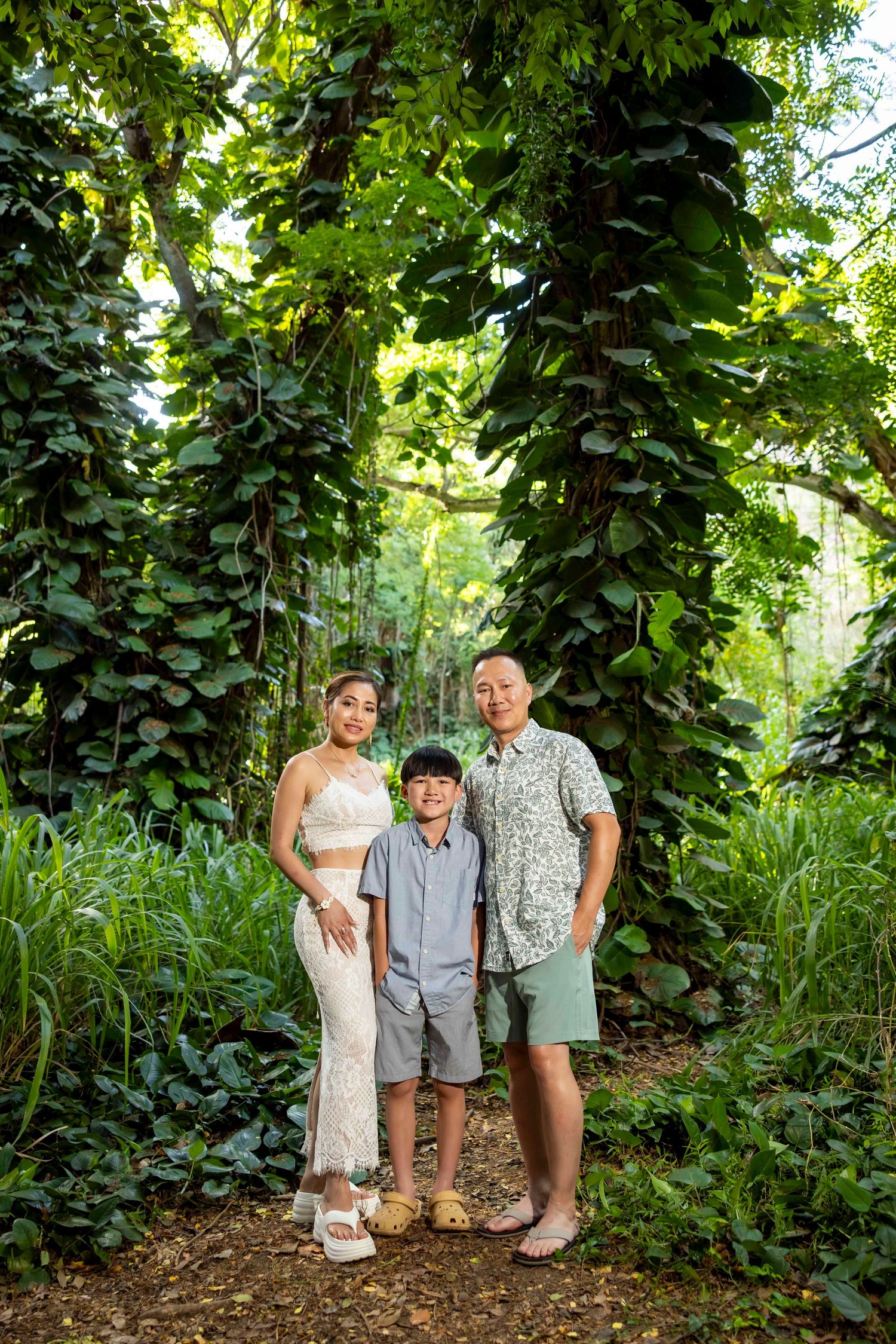 Smiling family of three poses on a dirt path in a lush tropical forest with vine-covered trees and tall green grasses.