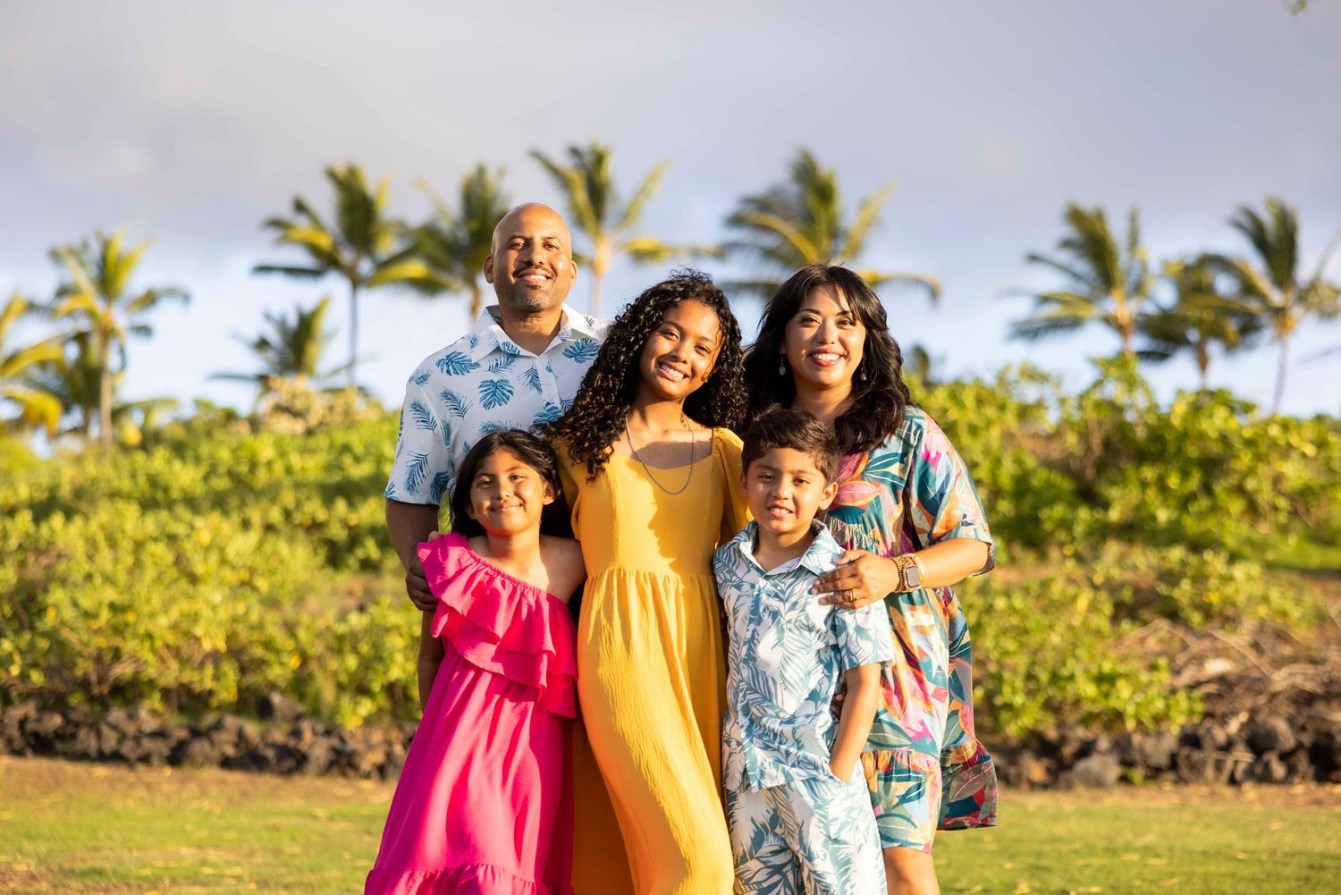 Smiling family of five in colorful tropical outfits posing together outdoors with palm trees and green shrubs in the background.
