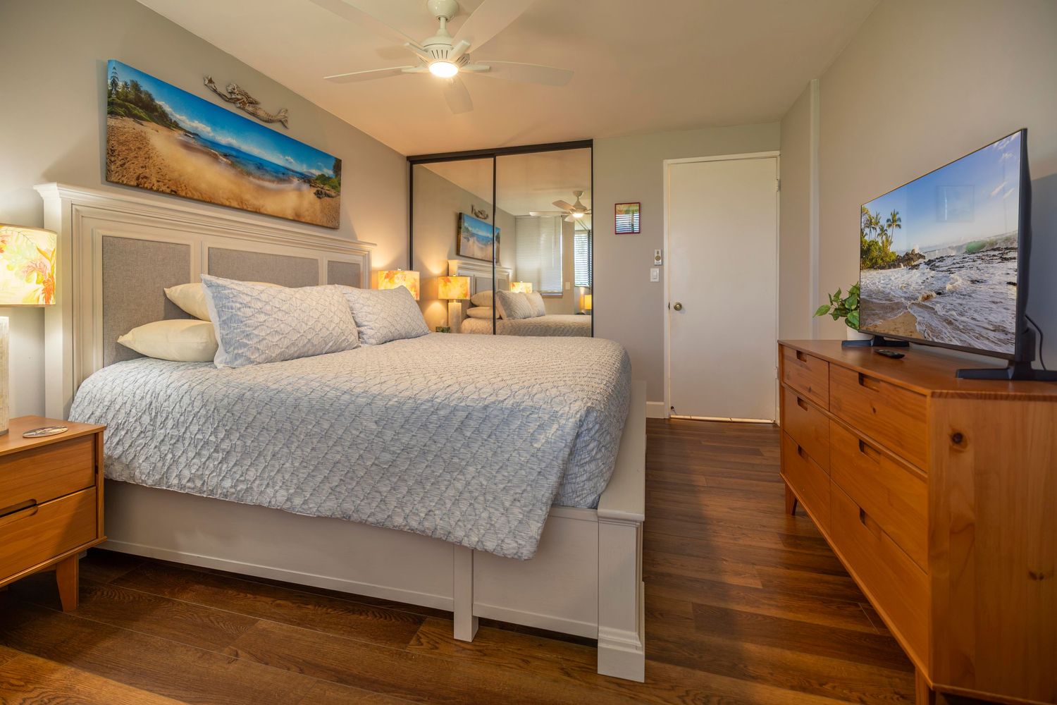 Bright bedroom with a king bed, tufted headboard, beach artwork, wood dresser with flat-screen TV, mirrored closet, and hardwood floors.