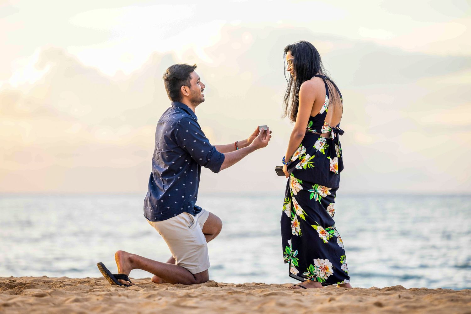 Man in a navy shirt kneels on a sandy beach at sunset, proposing to a woman in a floral dress beside the ocean. Makena