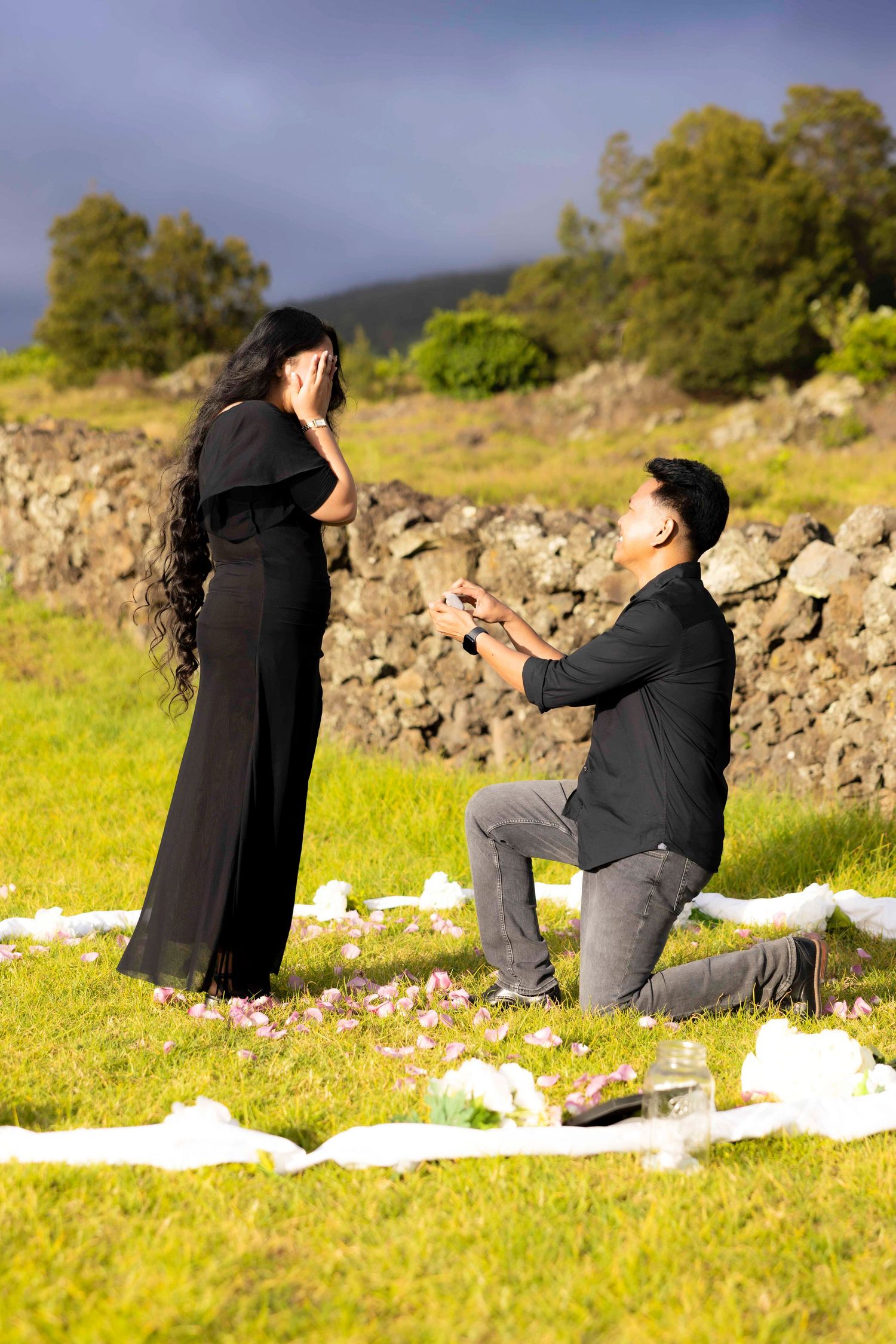 Man kneeling on grass proposes to a woman in a long black dress covering her face, with rose petals and a stone wall behind them. Upcountry