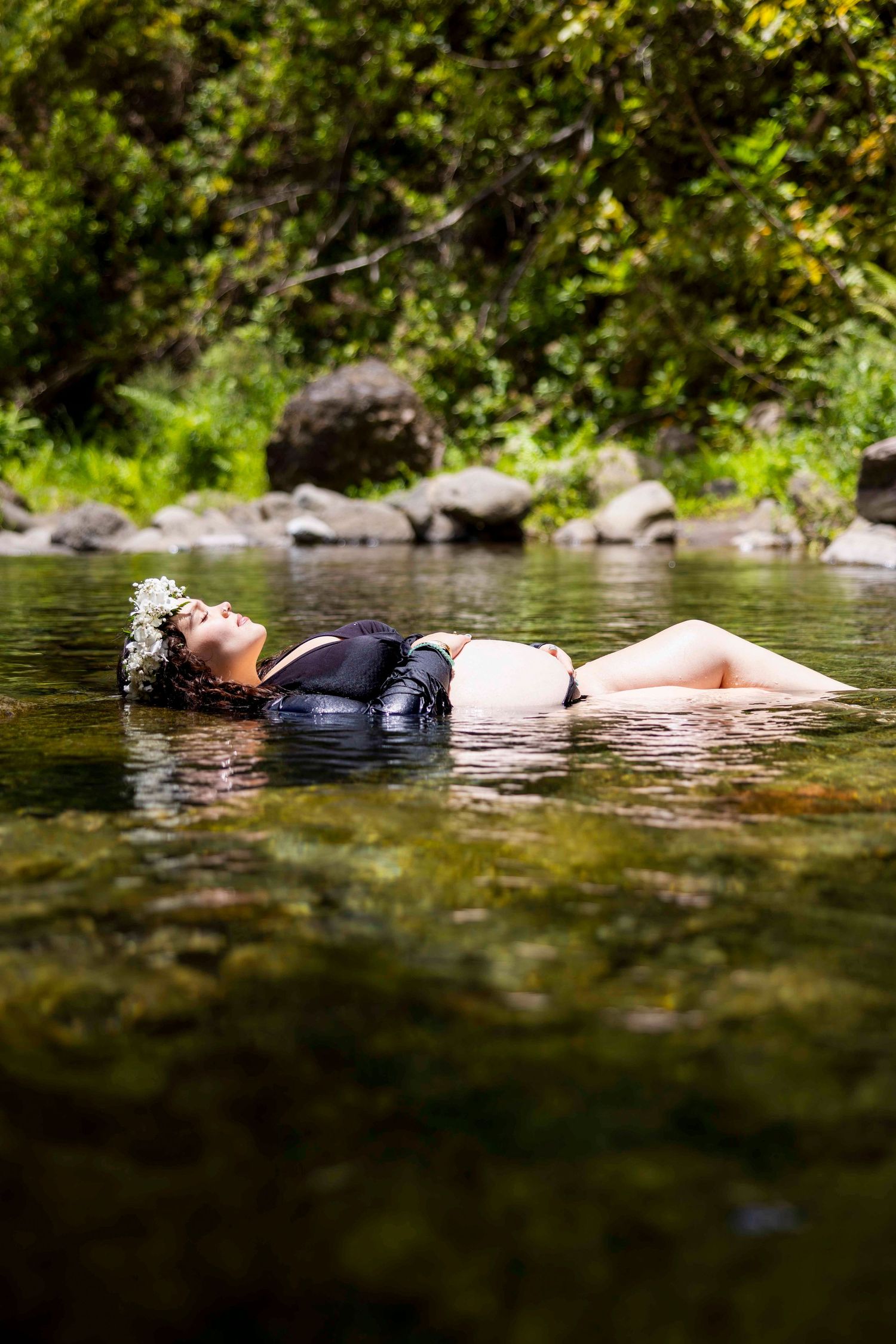 Woman wearing a white flower crown floats on her back in a clear shallow stream, surrounded by rocks and lush green foliage. Iao Valley