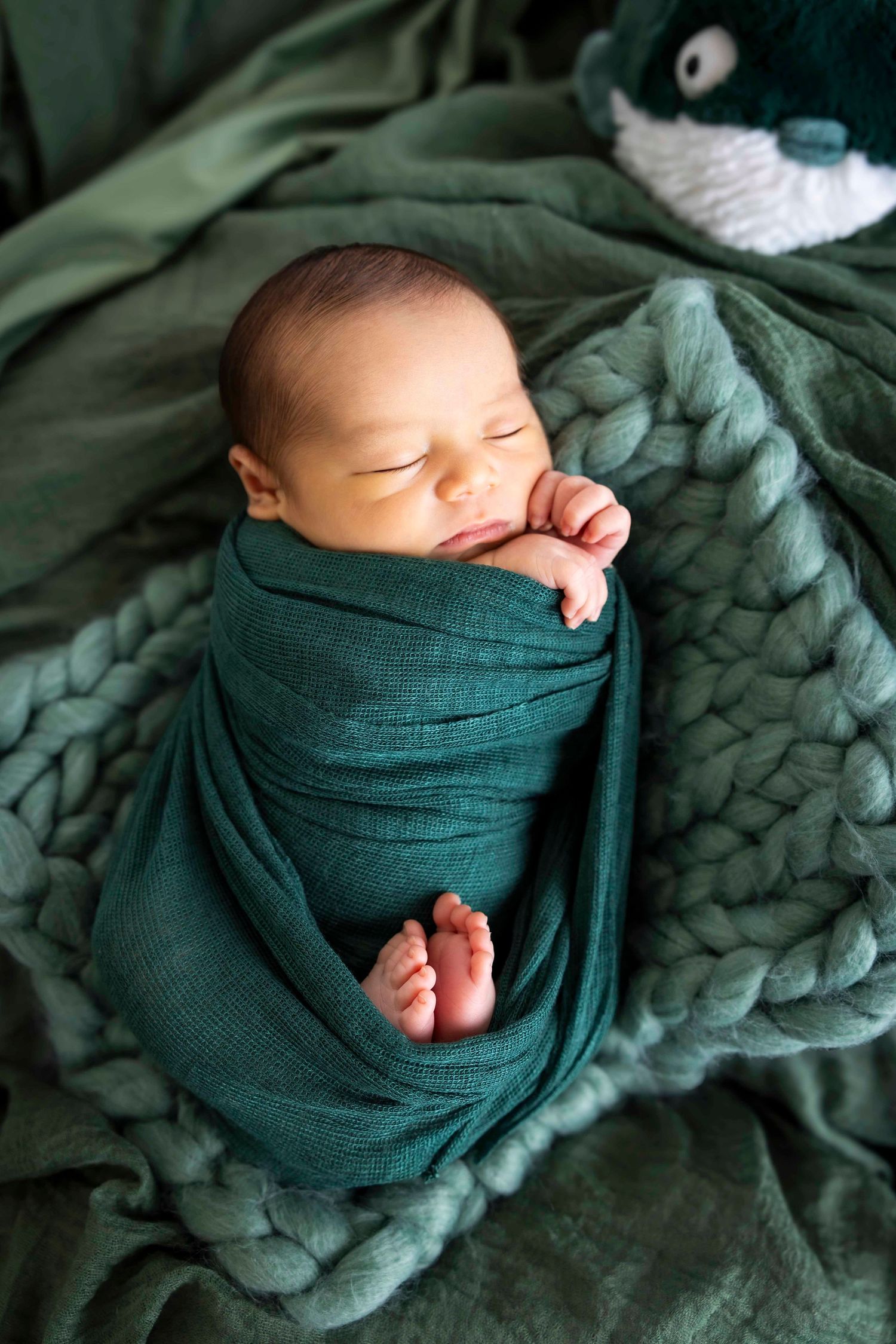 Sleeping newborn baby swaddled in a green wrap rests on a chunky knit blanket with tiny feet peeking out and a plush toy nearby.