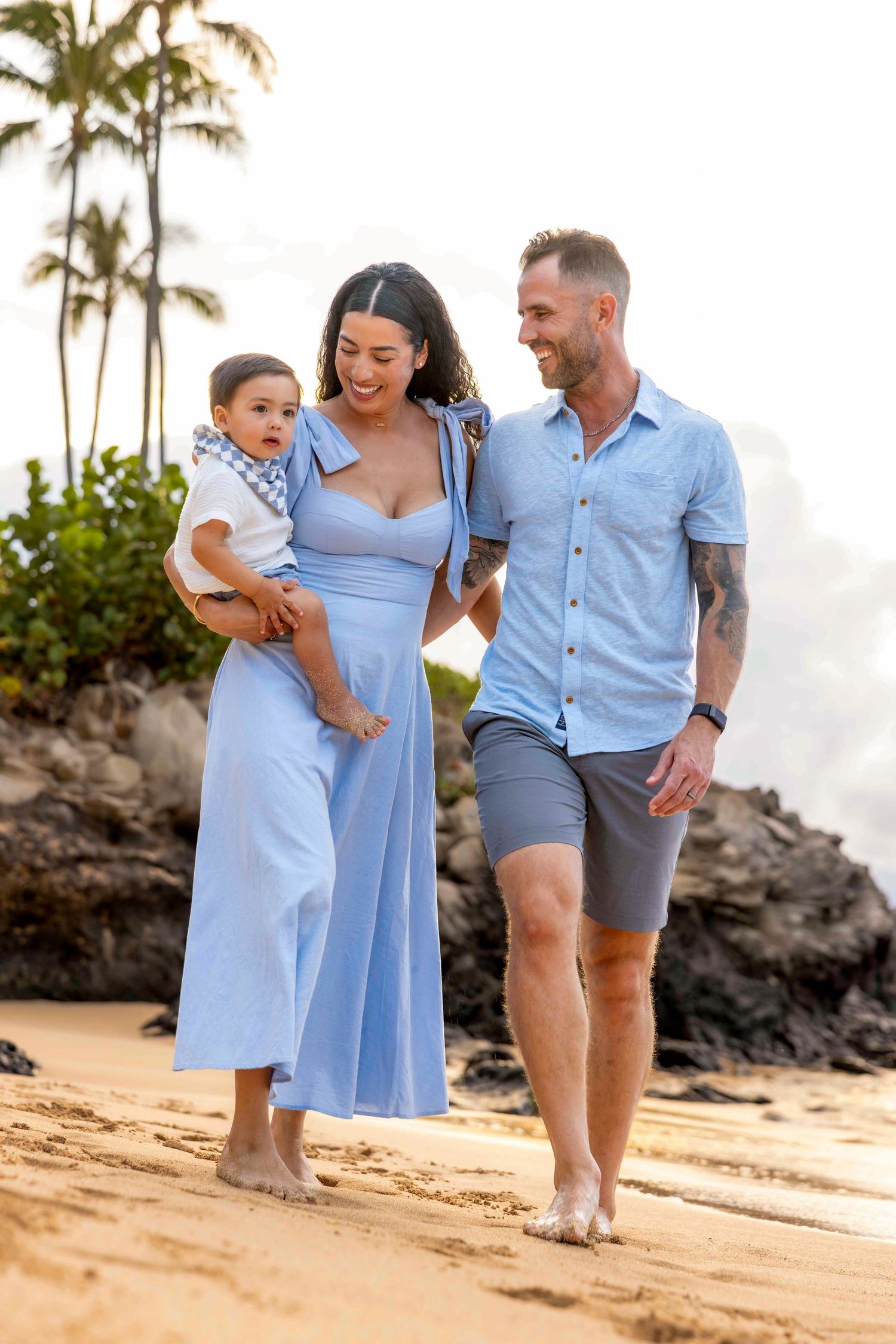 Smiling couple in matching light blue outfits stroll along a beach carrying their toddler, with rocky shoreline and greenery behind.