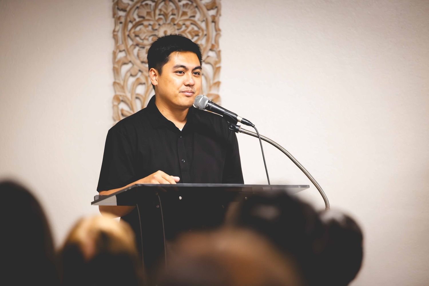 Man in a black shirt speaks into a microphone at a podium while an audience watches in the foreground of a room with carved wall decor.