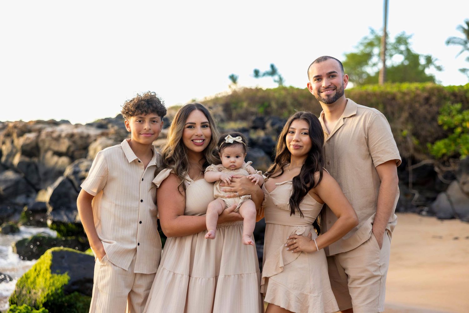 Family of six with a baby poses in neutral outfits on a Hawaii beach beside lava rocks and greenery for a portrait session.