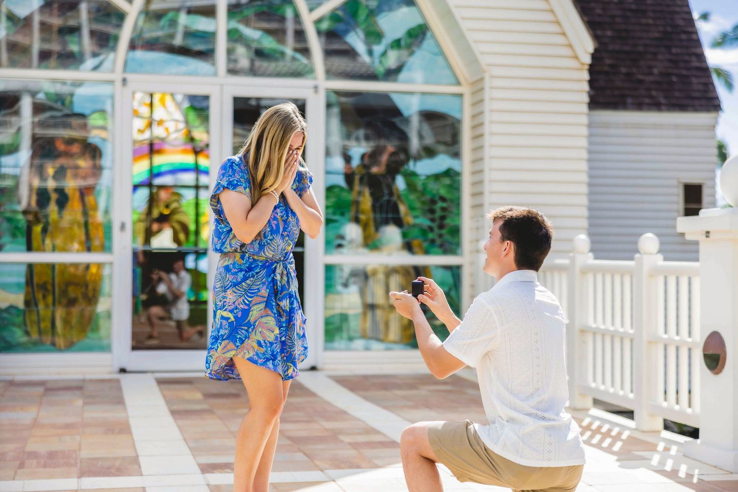 Man kneeling on one knee proposes to a surprised woman in a blue floral dress outside a chapel with stained glass windows. Wailea