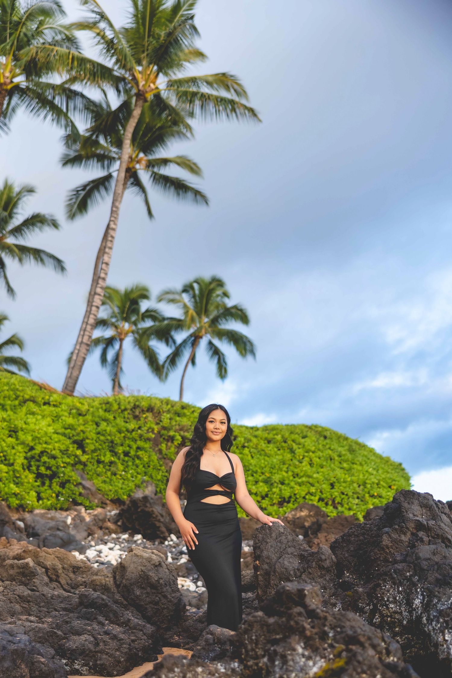 Woman in a black cutout dress posing on dark volcanic rocks with green shrubs and tall palm trees under a cloudy tropical sky. Makena