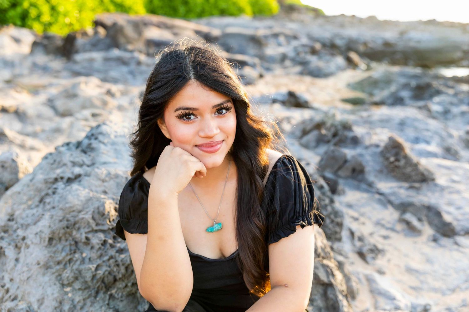 Young woman with long dark hair and a turquoise pendant necklace smiles while resting her chin on her hand on rocky coastal terrain. Napili