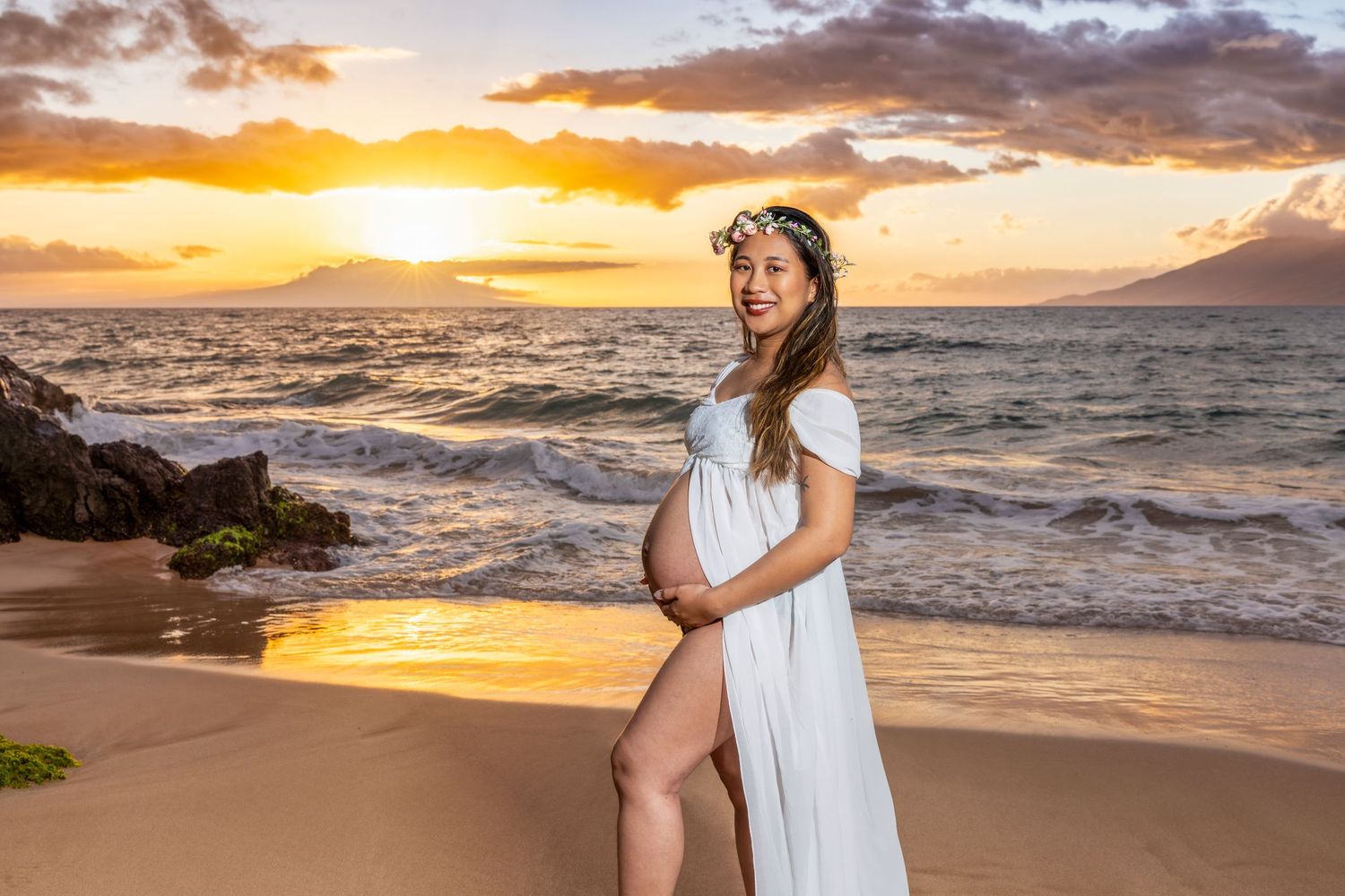 Pregnant woman in a flowing white dress and floral crown posing on a sandy beach at sunset with golden light over the ocean waves. Makena