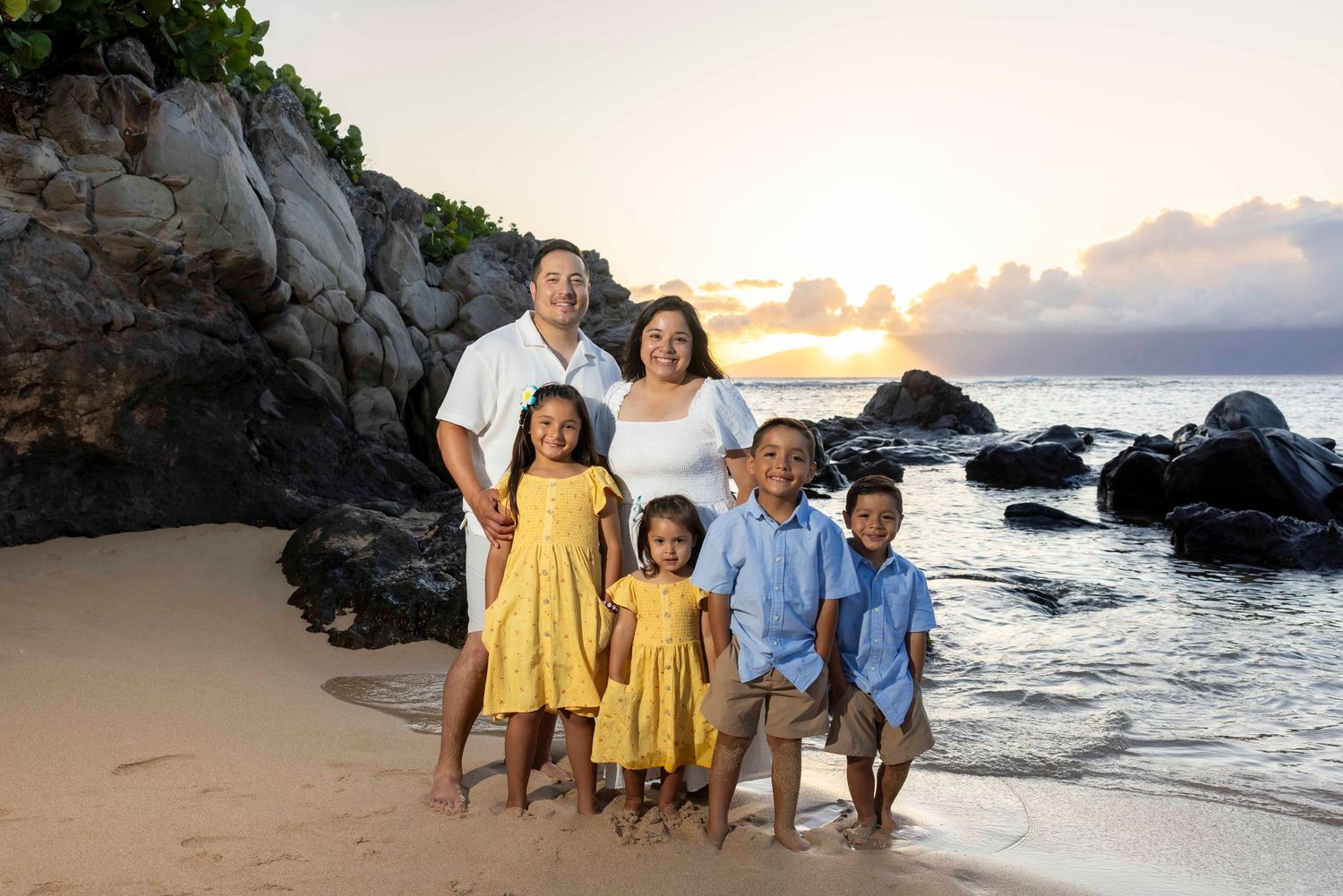 Family of six in coordinated outfits posing on a sandy beach at sunset with rocky lava formations and calm ocean waves behind them.
