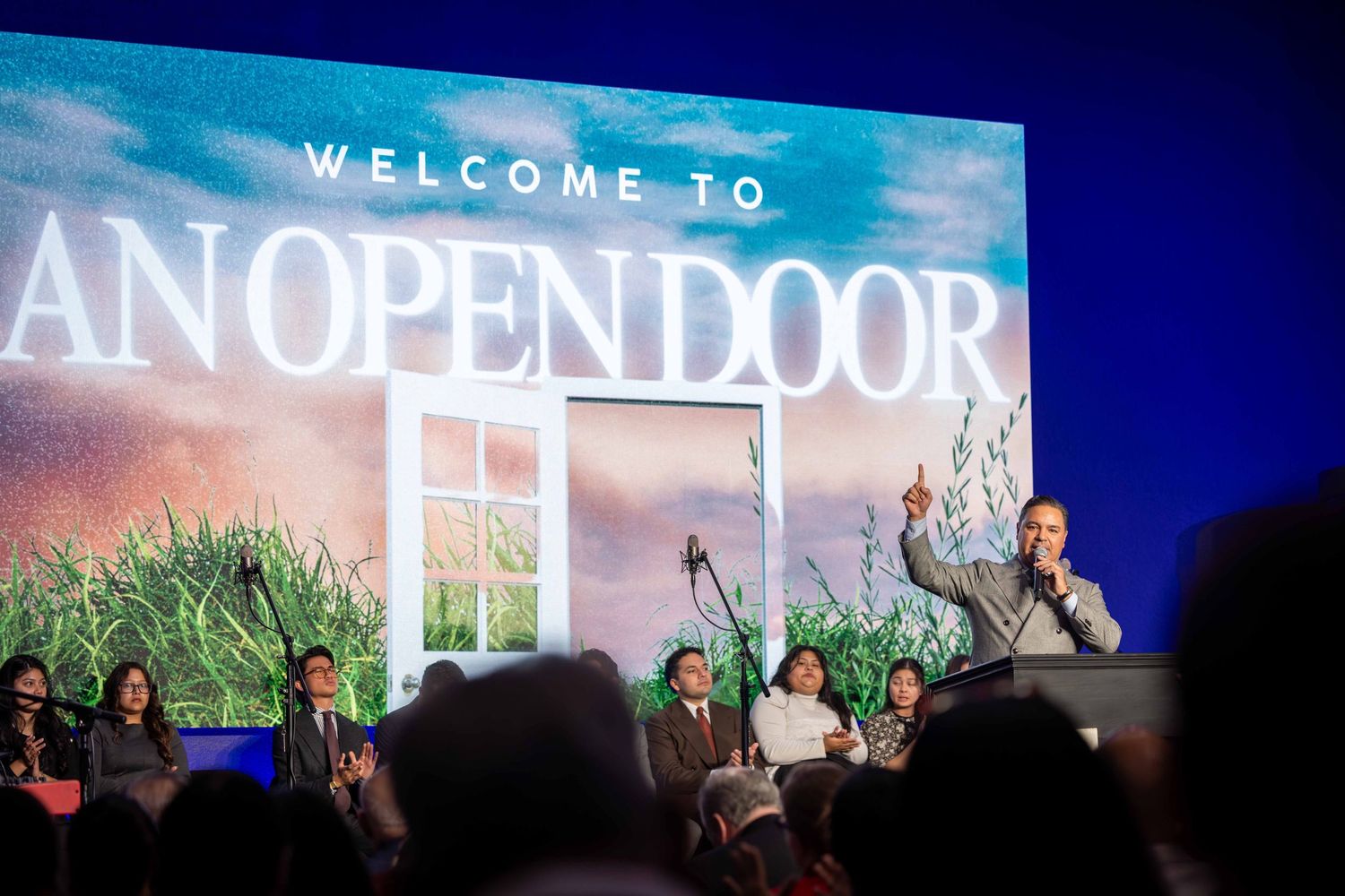 Speaker with raised finger addresses a seated audience on stage beneath a large Welcome to An Open Door screen showing an open window.