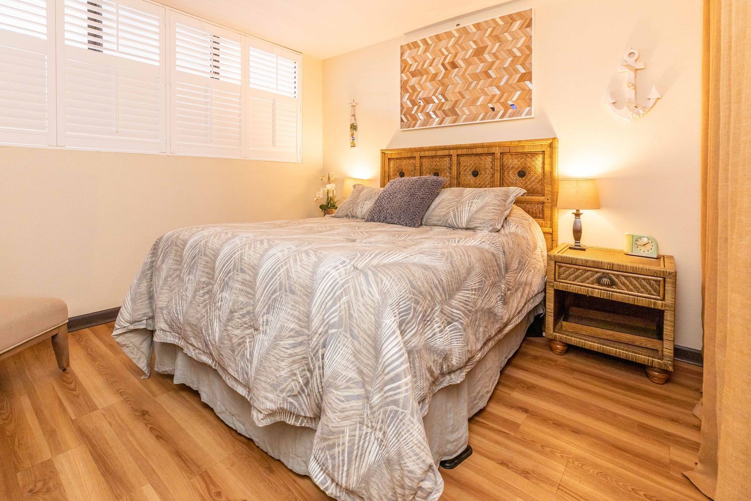 Cozy bedroom with a king bed, gray patterned comforter, rattan headboard, wood nightstand, plantation shutters and warm hardwood flooring.