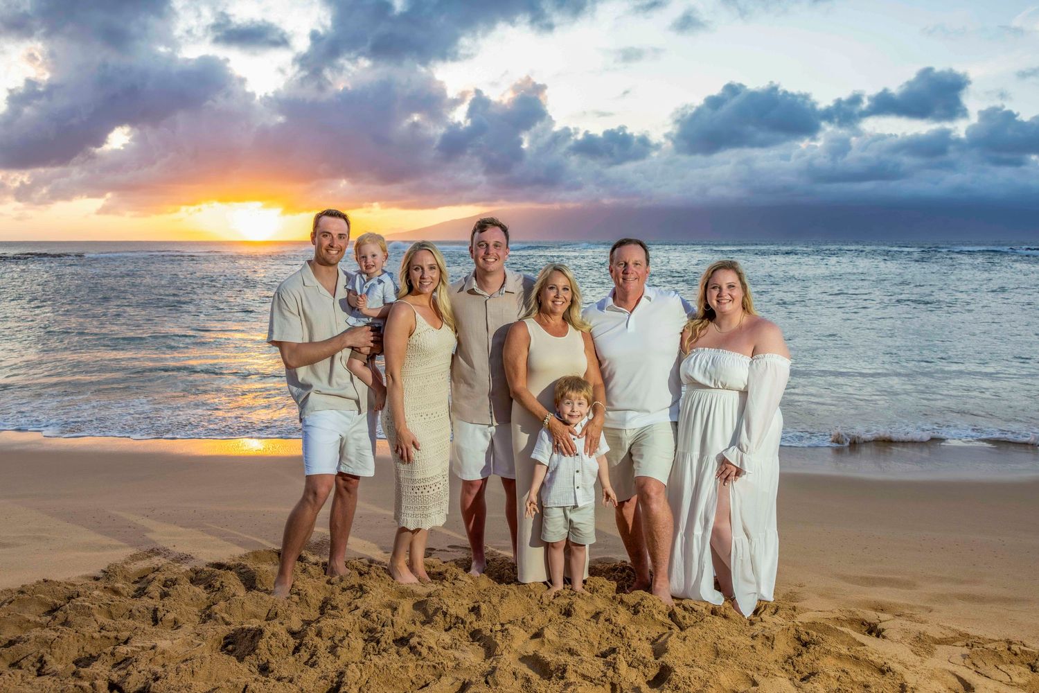 Extended family in white and neutral outfits poses together on a sandy beach at sunset with ocean waves and colorful clouds behind them.