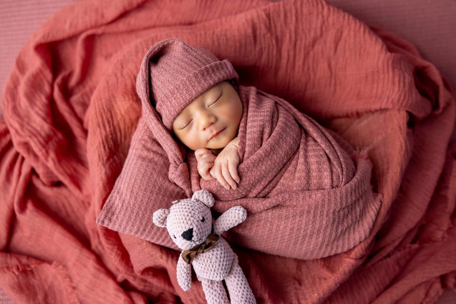 Sleeping newborn baby wrapped in a rose-colored blanket and matching knit hat rests beside a small crocheted teddy bear.