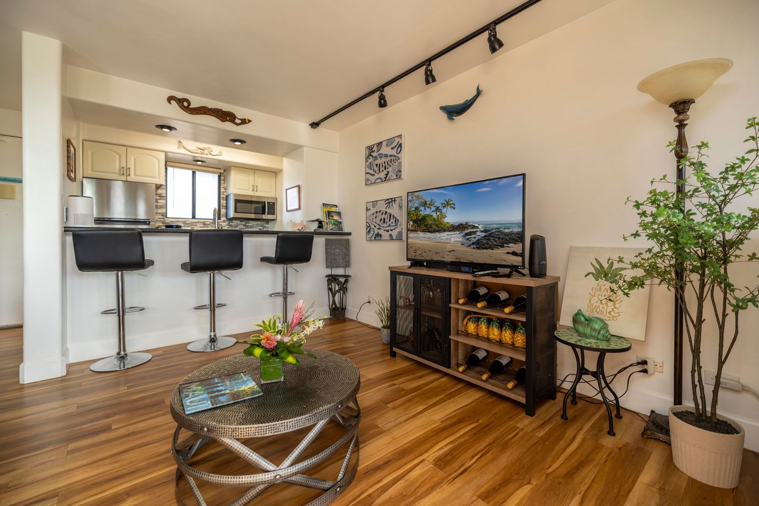 Bright open living area with hardwood floors, wall-mounted TV, round coffee table, potted plant, and kitchen breakfast bar with stools.