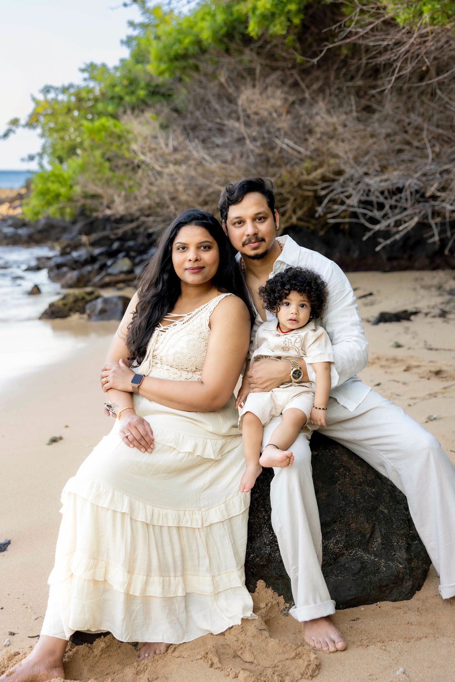 Parents in cream-colored outfits sit on a black lava rock with their curly-haired toddler on a tropical beach with green foliage behind.