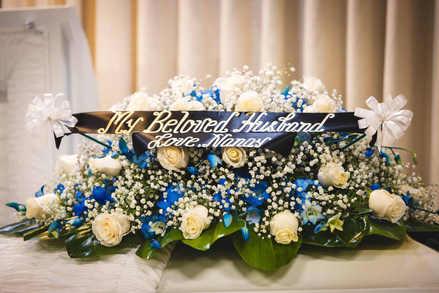 Funeral casket spray of white roses, blue flowers and baby's breath with a black sash reading My Beloved Husband, Love, Nancy.