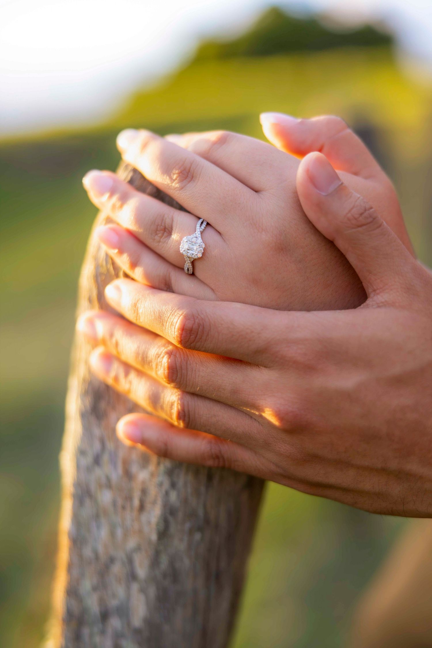 Couple's hands clasped atop a wooden fence post, showing a sparkling diamond engagement ring in warm golden sunset light. Upcountry