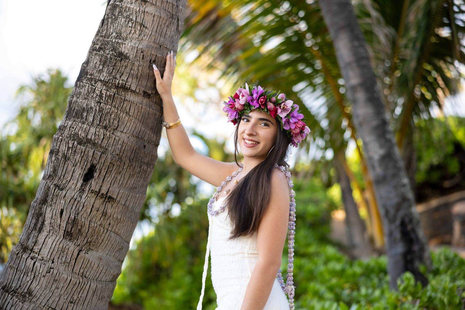 Smiling woman in a white dress and purple floral crown leans against a palm tree in a lush tropical setting. Paia