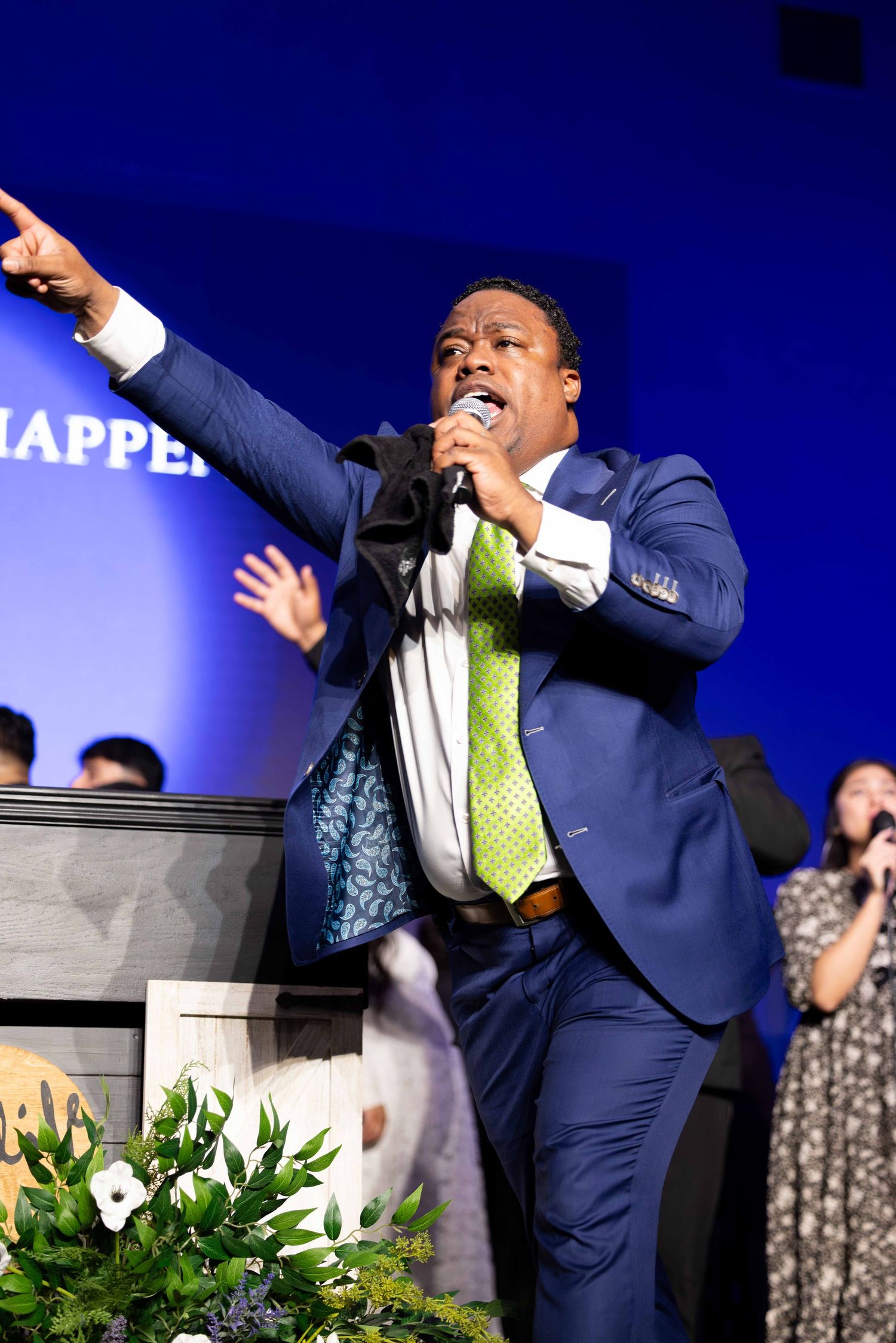 Man in a blue suit and green tie speaks passionately into a microphone, pointing outward on a stage with a blue backdrop and flowers.