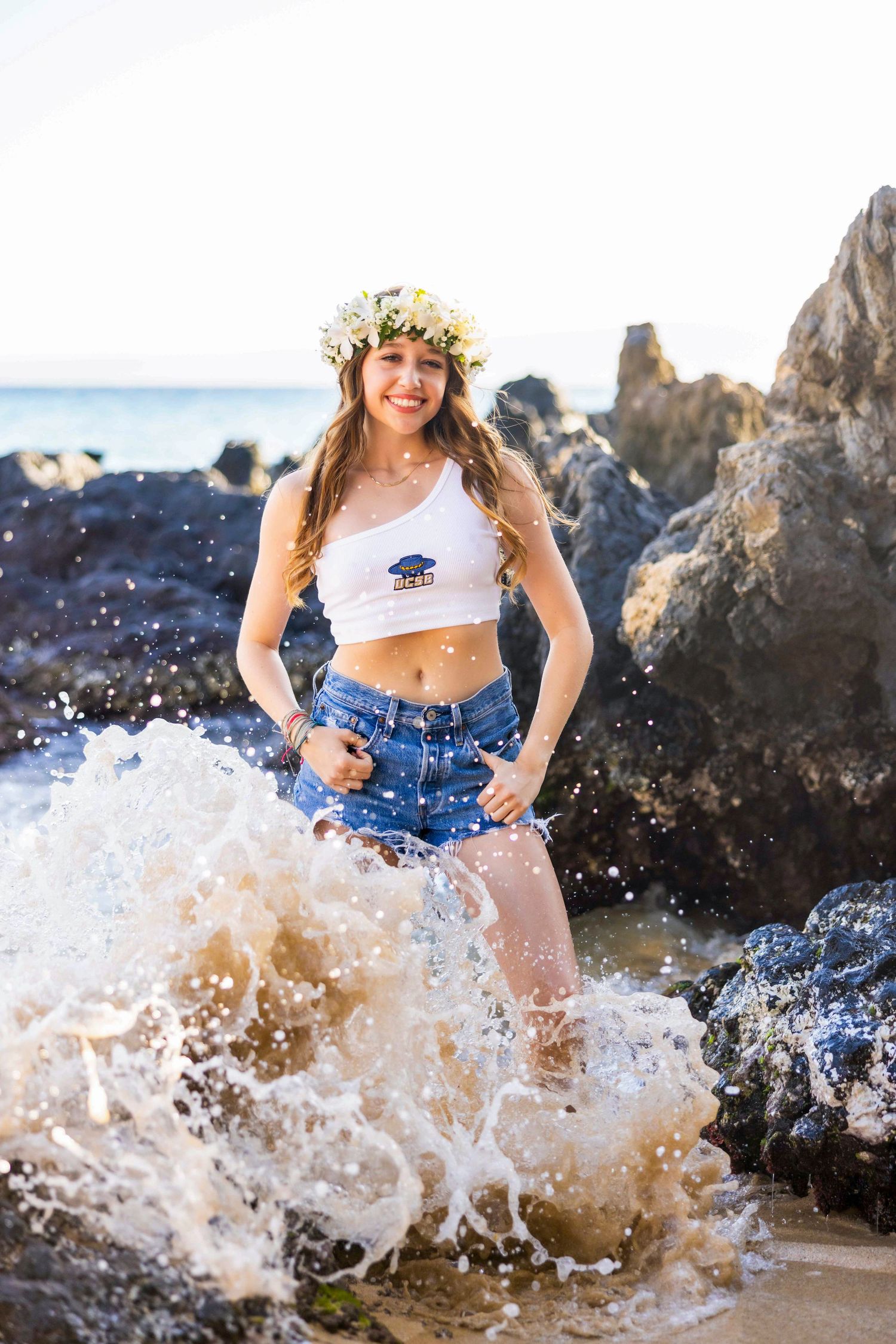 Smiling woman in a white crop top, denim shorts and flower crown stands on rocky shore as an ocean wave splashes around her. Makena