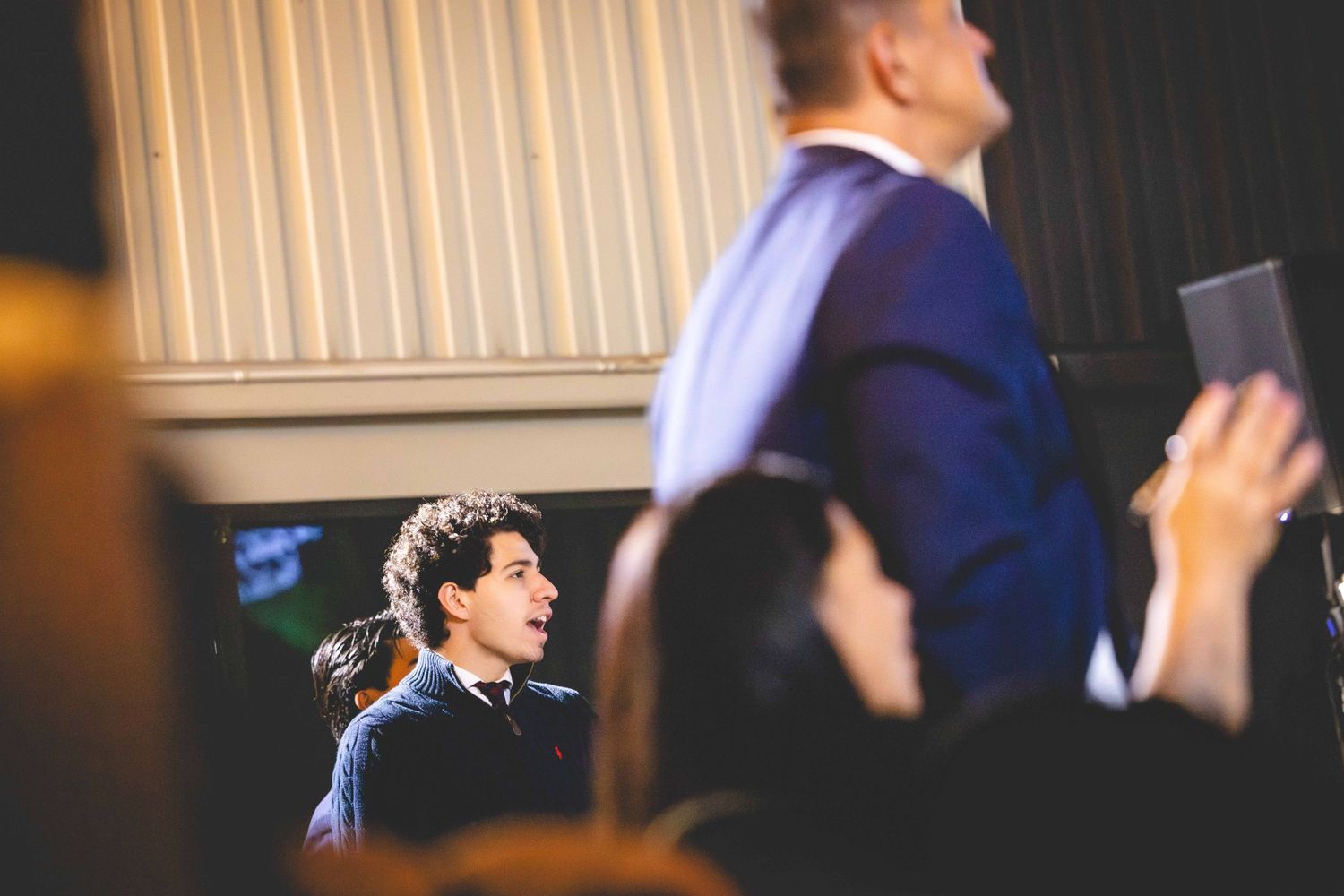 Young man with dark curly hair in a suit smiles from the audience while a blurred man in a blue jacket stands in the foreground at an event.