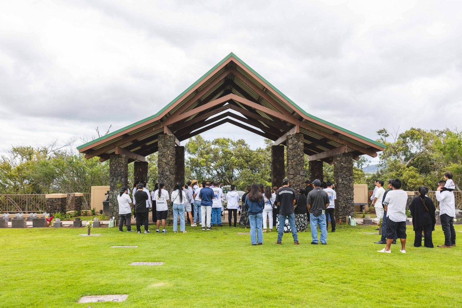 Group of people standing in a circle with heads bowed beneath a rustic wooden pavilion with a green roof on a lush lawn under cloudy skies.