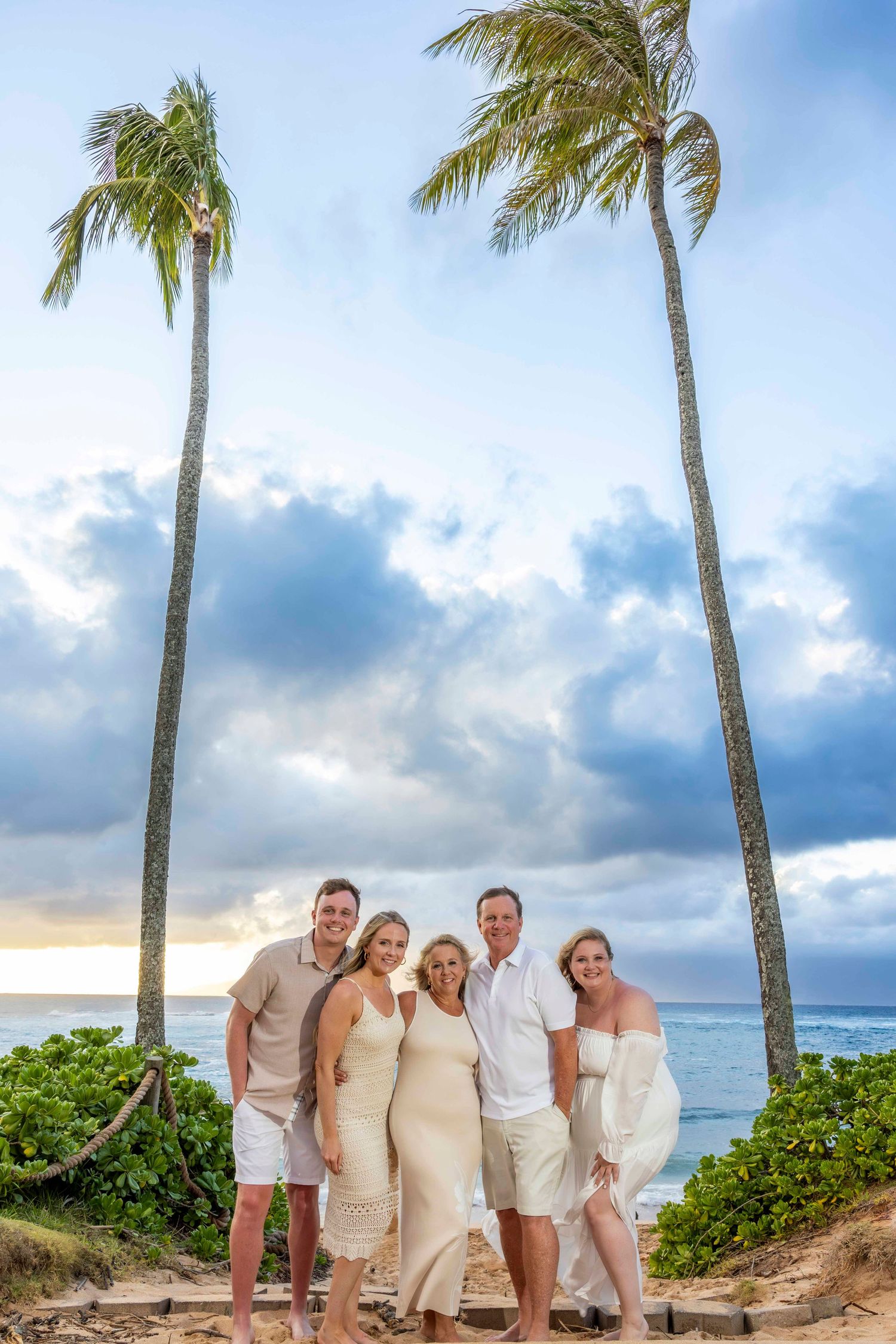 Family of five in neutral beach attire posing on a tropical shore between two tall palm trees at sunset with a cloudy sky. Kapalua