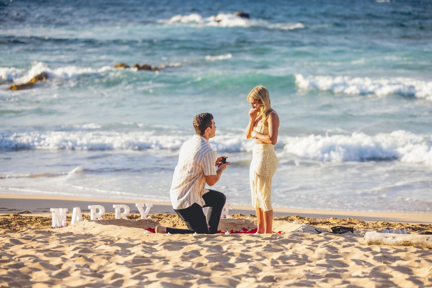 Man kneeling on a sandy beach proposing to a smiling woman with ocean waves behind them and decorative letters spread on the sand. Iroonwood