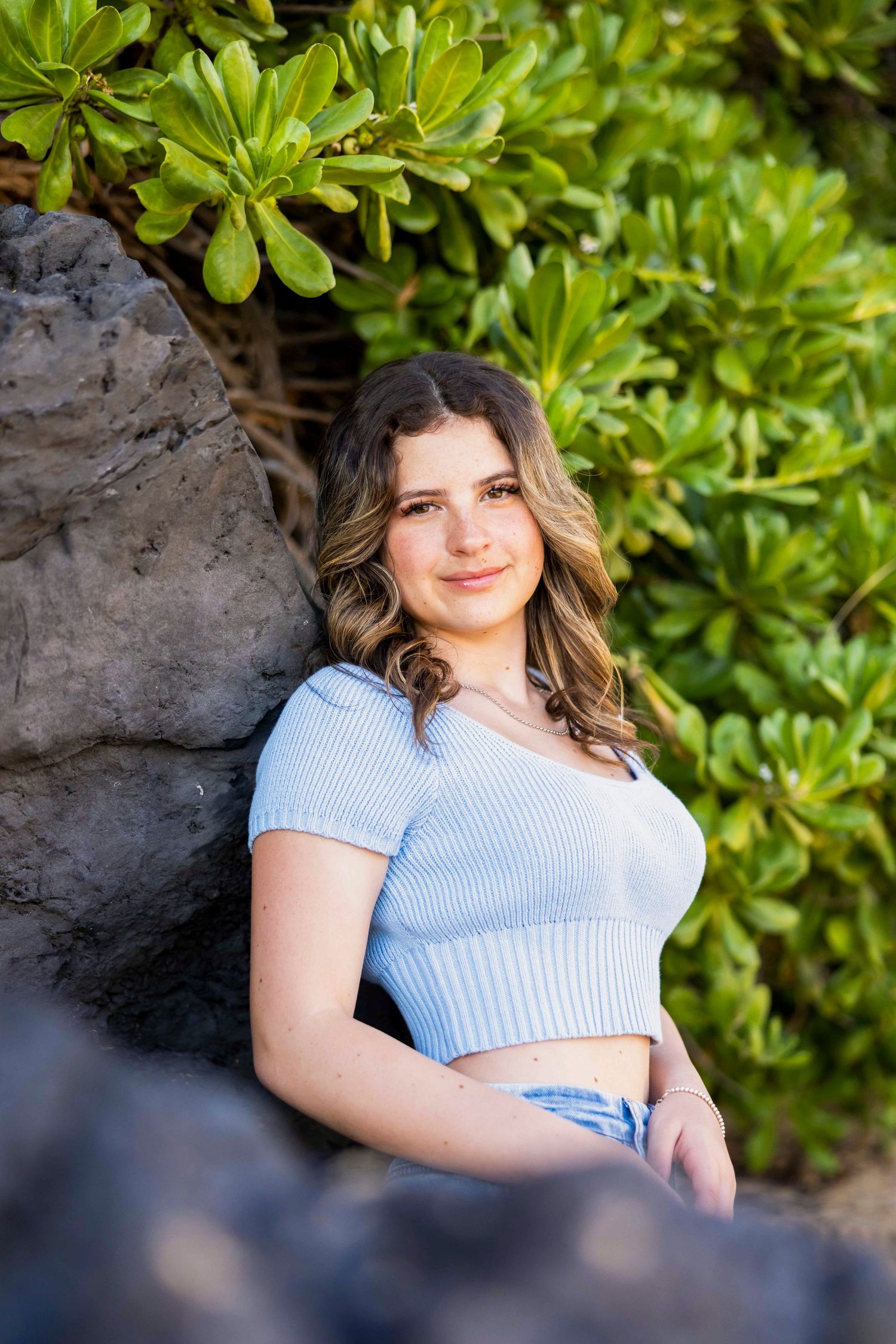 Young woman with wavy brown hair in a light blue ribbed crop top leans against a dark lava rock beside lush green tropical foliage. Makena