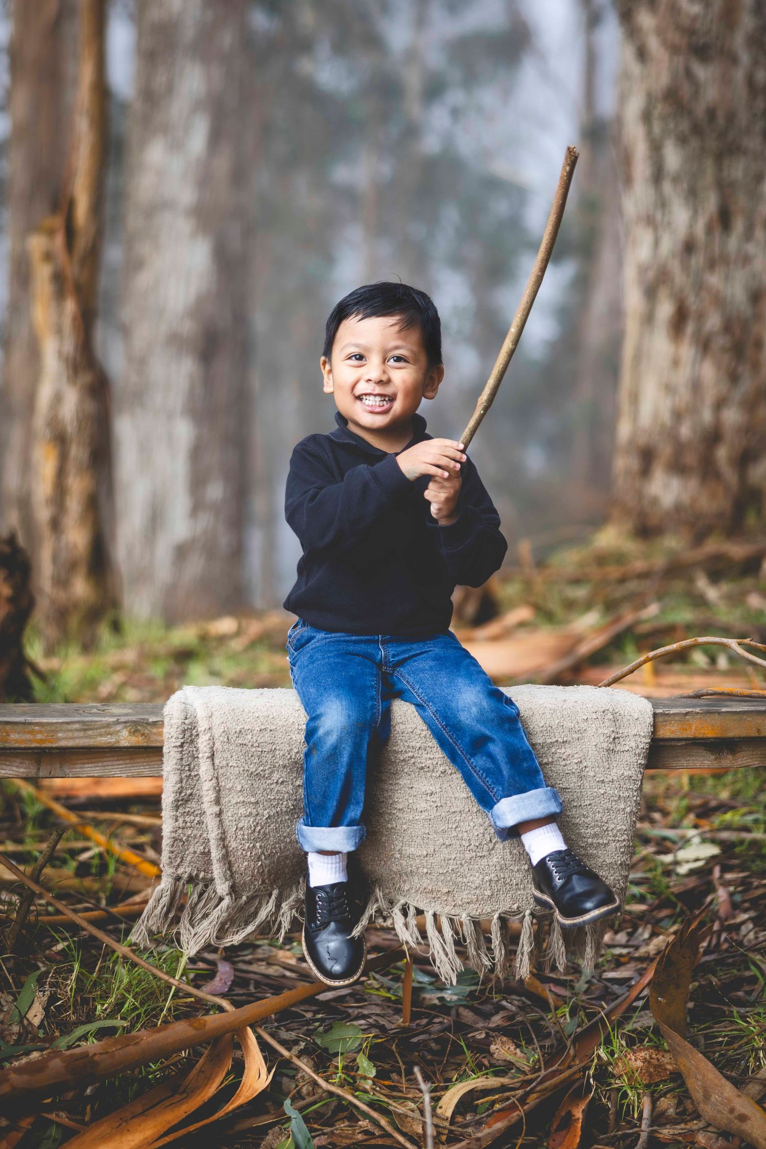 Smiling young boy in a navy sweater and jeans sits on a wooden bench with a woven blanket, holding a stick in a misty eucalyptus forest.