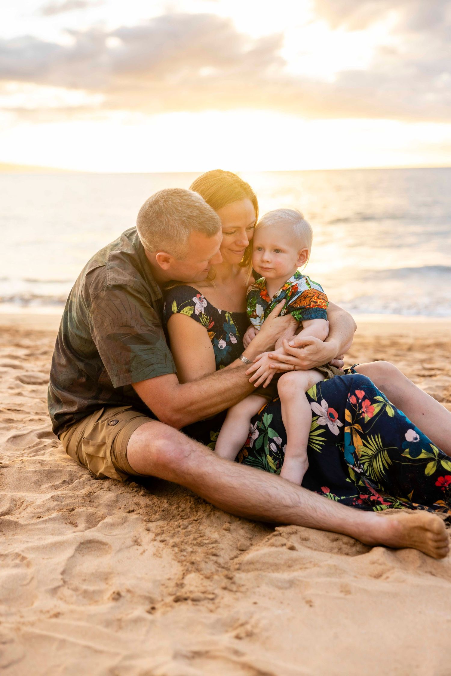 Family of three in tropical-print outfits cuddling on a sandy beach at sunset, with a toddler smiling as gentle ocean waves roll in behind.