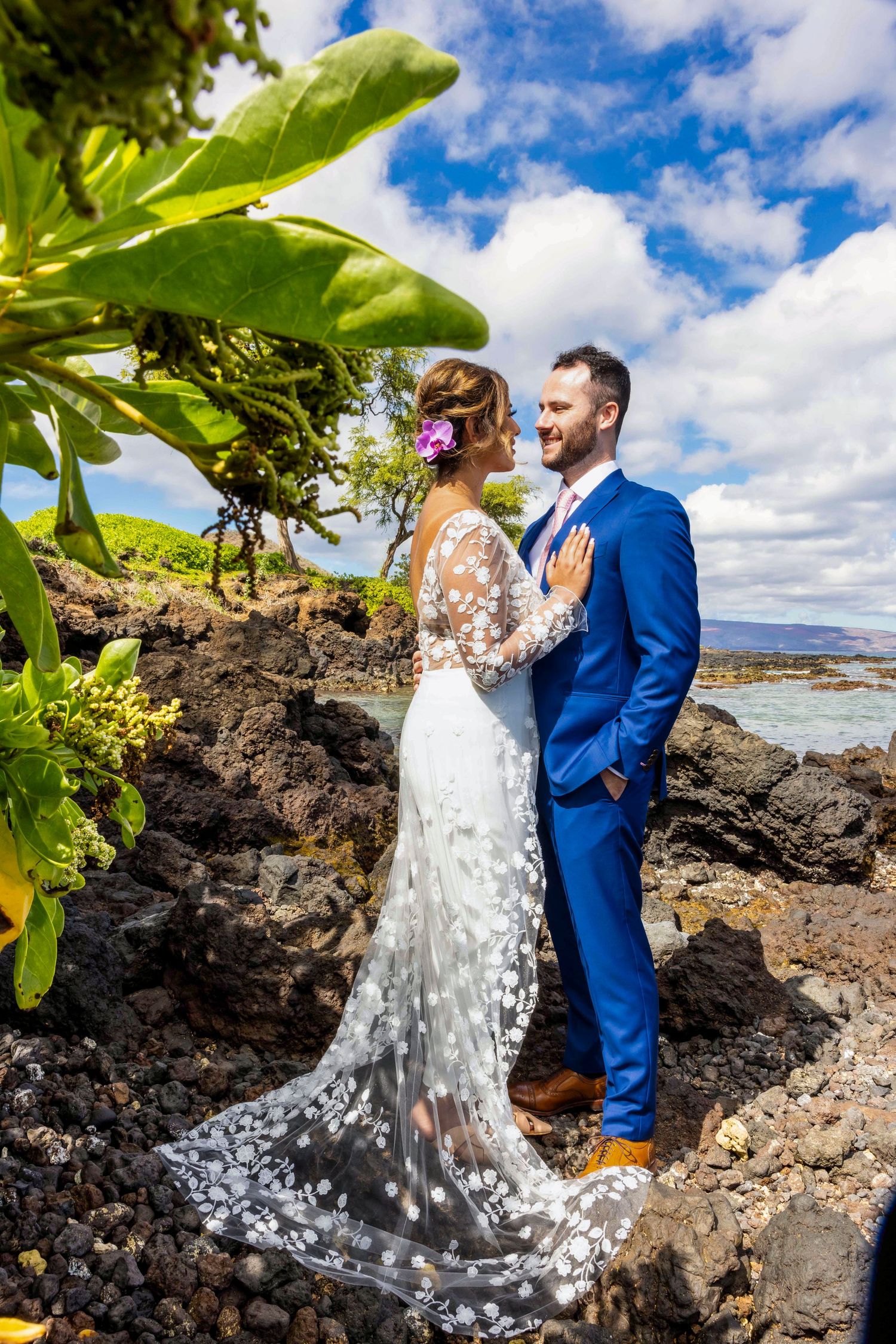 Bride in a lace floral wedding gown and groom in a blue suit pose on volcanic rocks beside the ocean at a tropical beach wedding. Makena